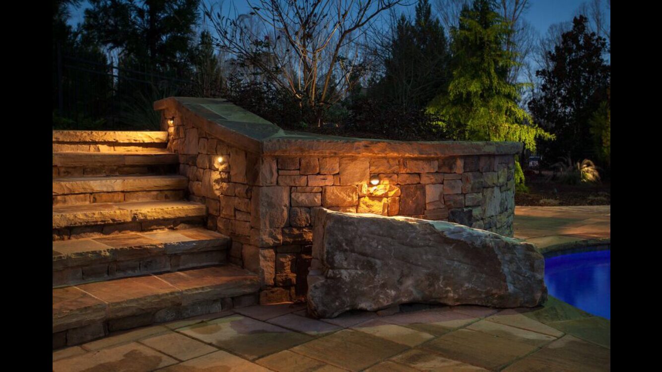 Stacked-stone retaining wall and steps lit with recessed fixtures beside a pool.